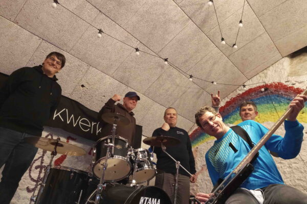 5 Mitglieder der Band auf der Bühne, von unten fotografiert, lächeln, zwei ballen die Faust, in der Mitte ein Schlagzeug, einer hält eine E-Gitarre