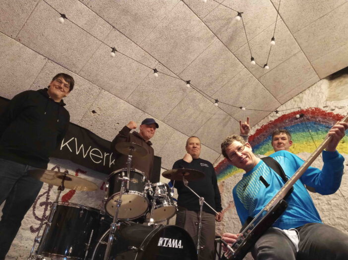 5 Mitglieder der Band auf der Bühne, von unten fotografiert, lächeln, zwei ballen die Faust, in der Mitte ein Schlagzeug, einer hält eine E-Gitarre