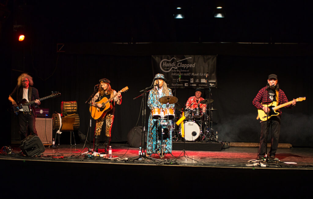 5 Mitglieder der Band beim Auftritt live auf der Bühne, mit Schlagzeug, Gitarre, E-Bass, E-Gitarre und Congas. Die Sängerin trägt ein auffälliges Mantelartiges blaues Gewandt