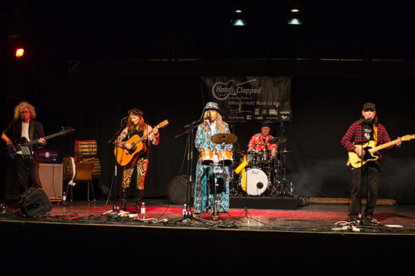 5 Mitglieder der Band beim Auftritt live auf der Bühne, mit Schlagzeug, Gitarre, E-Bass, E-Gitarre und Congas. Die Sängerin trägt ein auffälliges Mantelartiges blaues Gewandt
