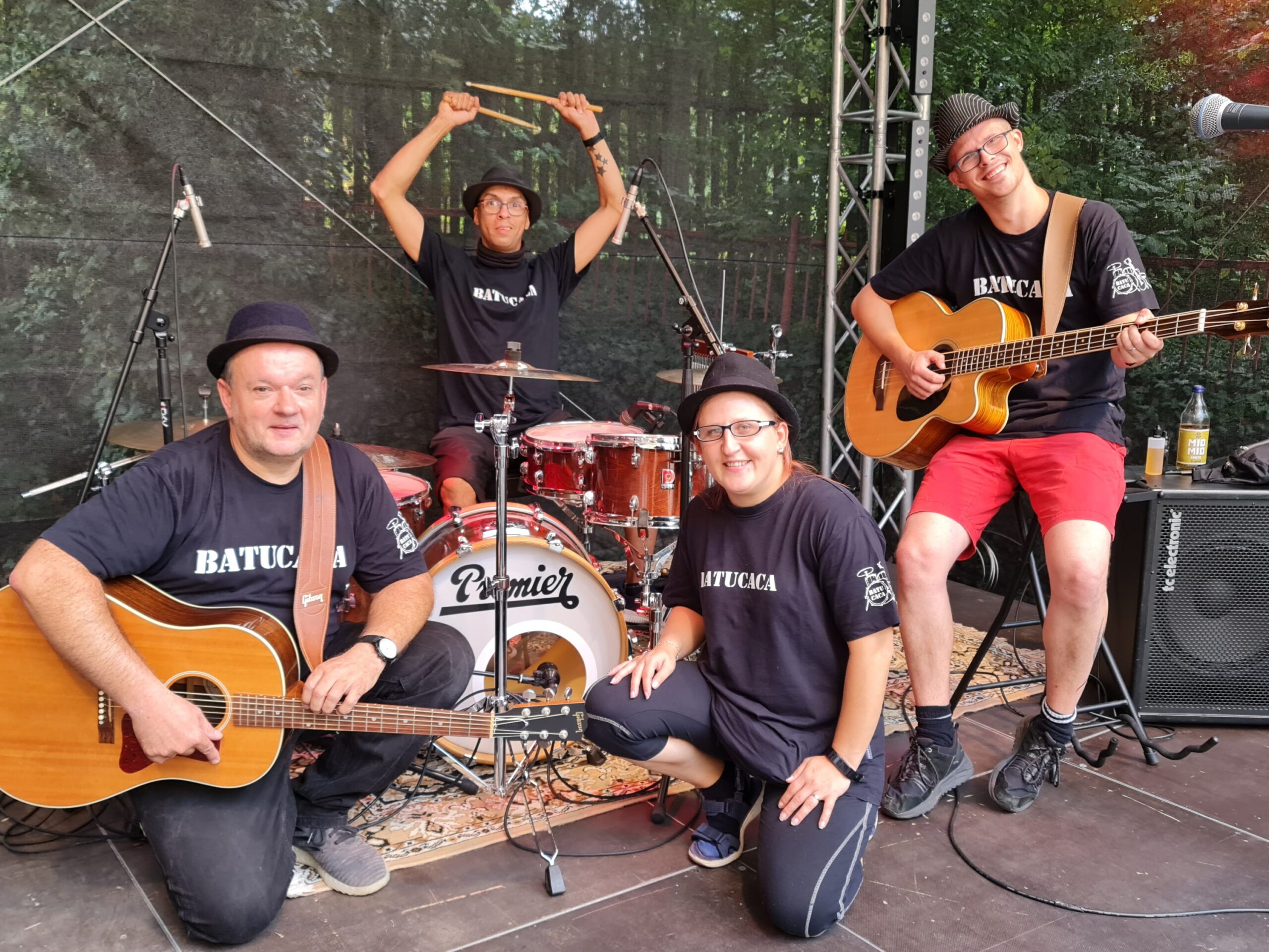 4 Mitglieder der Band auf einer Bühne im Grünen zum Gruppenfoto mit Schlagzeug und zwei Gitarren gruppiert.