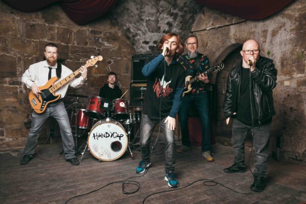 5 Mitglieder der Band in einem Kellergewölbe, 3 spielen Ihre Instrumente, Gitarre, Schlagzeugg, E-Bass und zwei Sänger mit Mikros