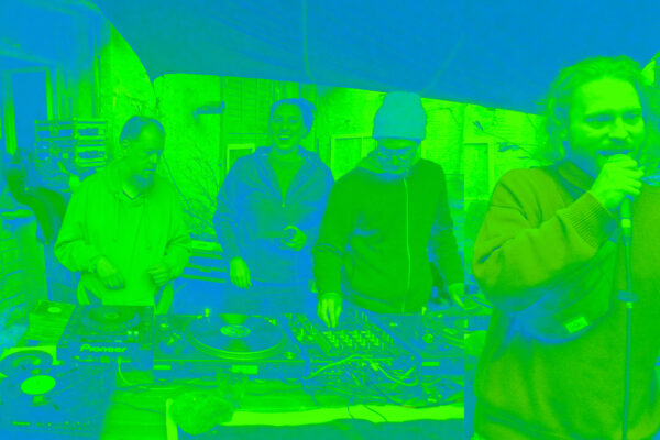 4 Mitglieder des DJ-Kollektivs, einer mit Mikro vor und drei hinter einem Tisch mit Plattentellern und Mixer. Das ganze Bild ist grün eingefärbt.