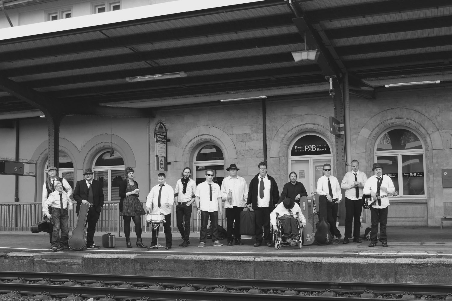 Schwarz weiss Foto von 14 Mitgliedern der Band auf einem Bahnsteig, fast alle mit weißen Hemden und Schlipsen, einer im Rollstuhl, einige mit Instrumentenkoffern.