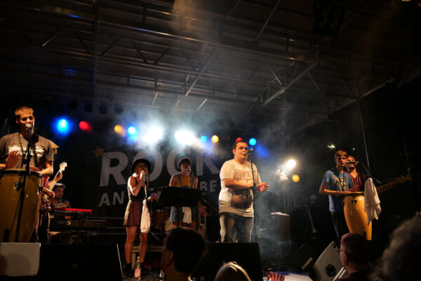 6 Mitglieder der Band live beim Auftritt auf einer Bühne (innen) mit bunten Lichtern und Bühnennebel.