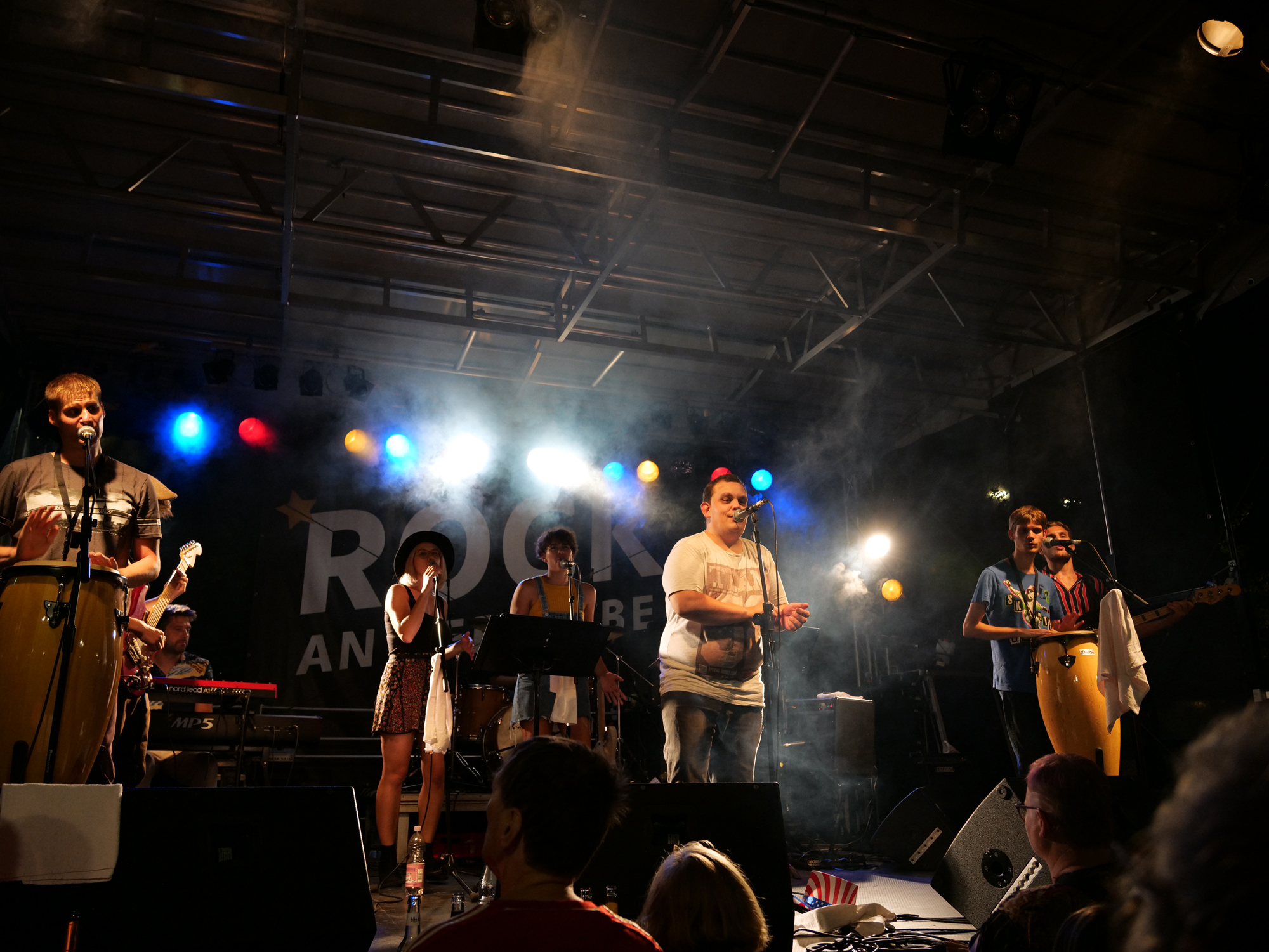 6 Mitglieder der Band live beim Auftritt auf einer Bühne (innen) mit bunten Lichtern und Bühnennebel.