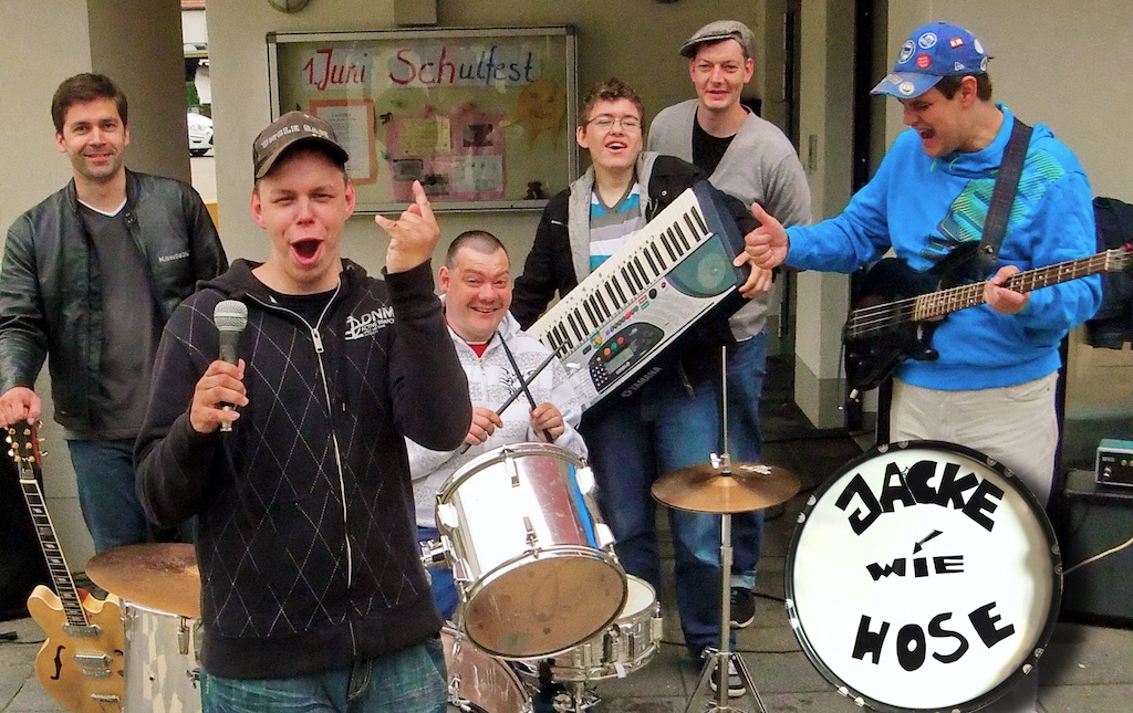 6 Mitglieder der Band gut glelaunt vor einem Gebäude mit Instrumenten (Schlagzeug, Mikro, Gitarre, Keyboard)