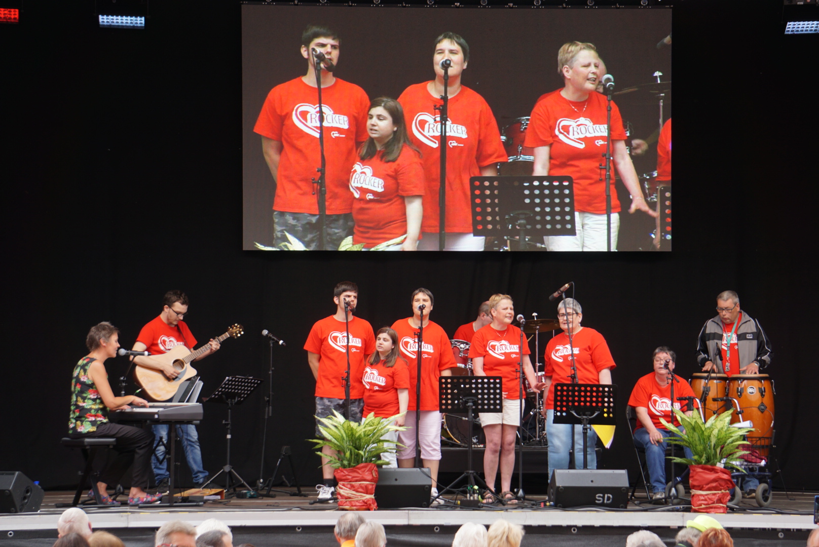 10 Mitglieder, bis auf eine am Keyboard alle in roten T-Shirts auf einer Außenbühne beim Auftritt.