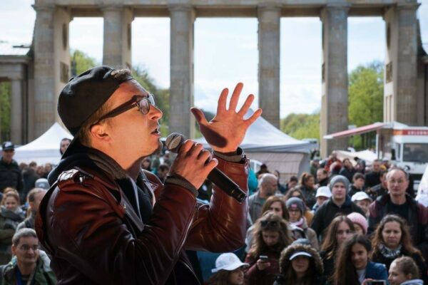 Ein Sänger mit Basecap singt ins Mikro, im Hintergrund das Brandenburger Tor.