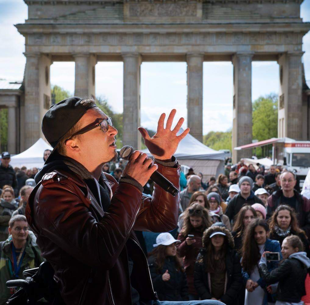 Ein Sänger mit Basecap singt ins Mikro, im Hintergrund das Brandenburger Tor.