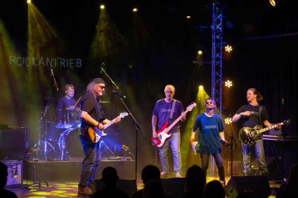 Buntfoto von fünf Musiker, die live auf der Bühne stehen, mit Schlagzeig, E-Gitarre, zwei Bässen und einer Sängerin. Dazu dunle Bühnenbeleuchtung und im Hintergrund Schriftzug "RockAntrieb"