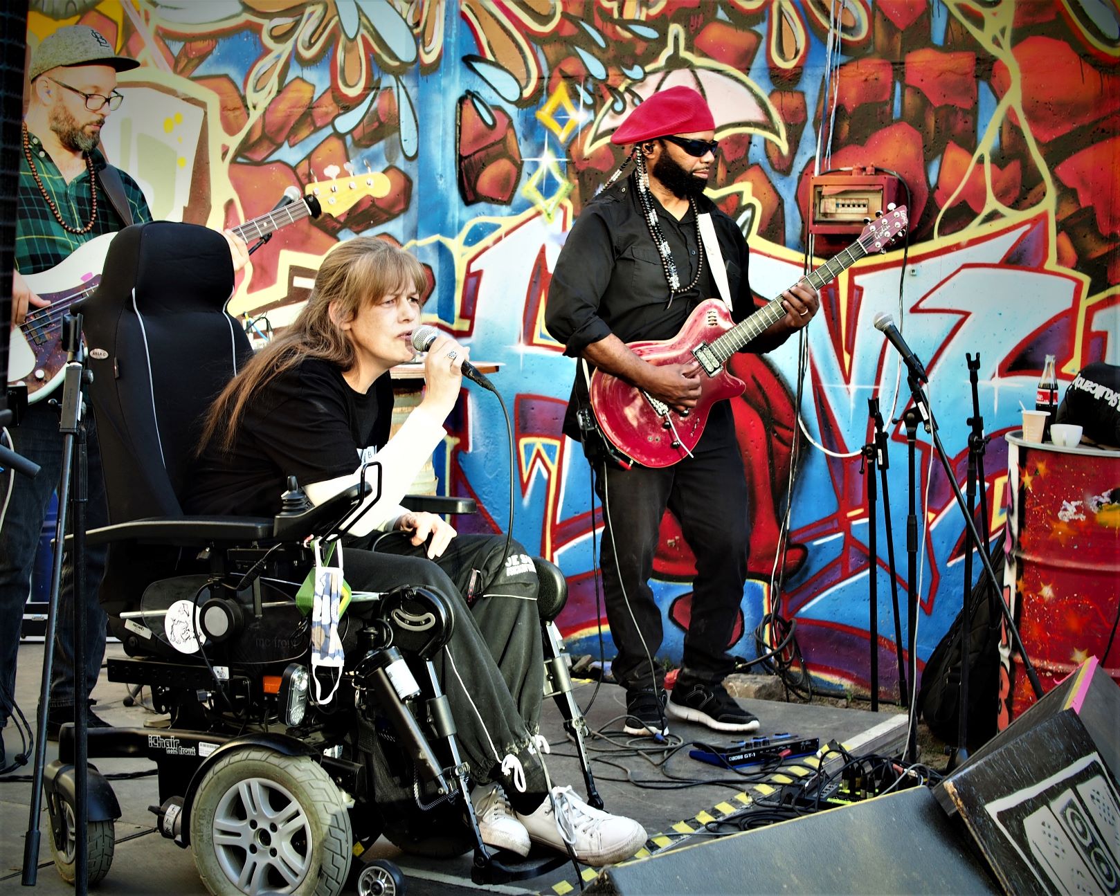 Claudia Russo (dat Muddi) live singend auf einer Freilichtbühne vor einer bunten Graffiti Wand, im Rollstuhl sitzend