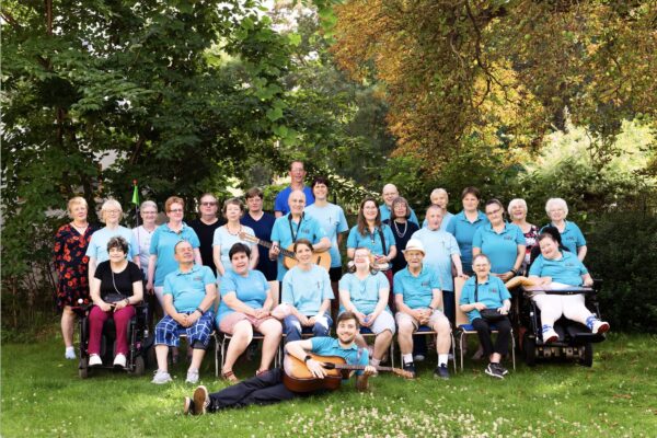 Bundfoto von 29 Mitgliedern eines Chores, unterschiedlichsten Alters, die meisten mit einem hellblauen T-Shirt bekleidet. Sie sitezen und stehen in zwei Reihen und gucken freundliche in die Kamera. Ein Mitglied liegt davor auf der Weise, mit einer Gitarre in der Hand. Rundherum grün von wiese und Bäumen.