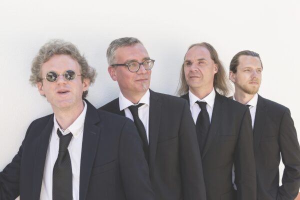 Foto der Band Blind Foundation. Die 4 Musiker stehen lächelnd vor einer weißen Wand. Sie tragen weiße Hemden und schwarze Anzüge mit schwarzen Krawatten.