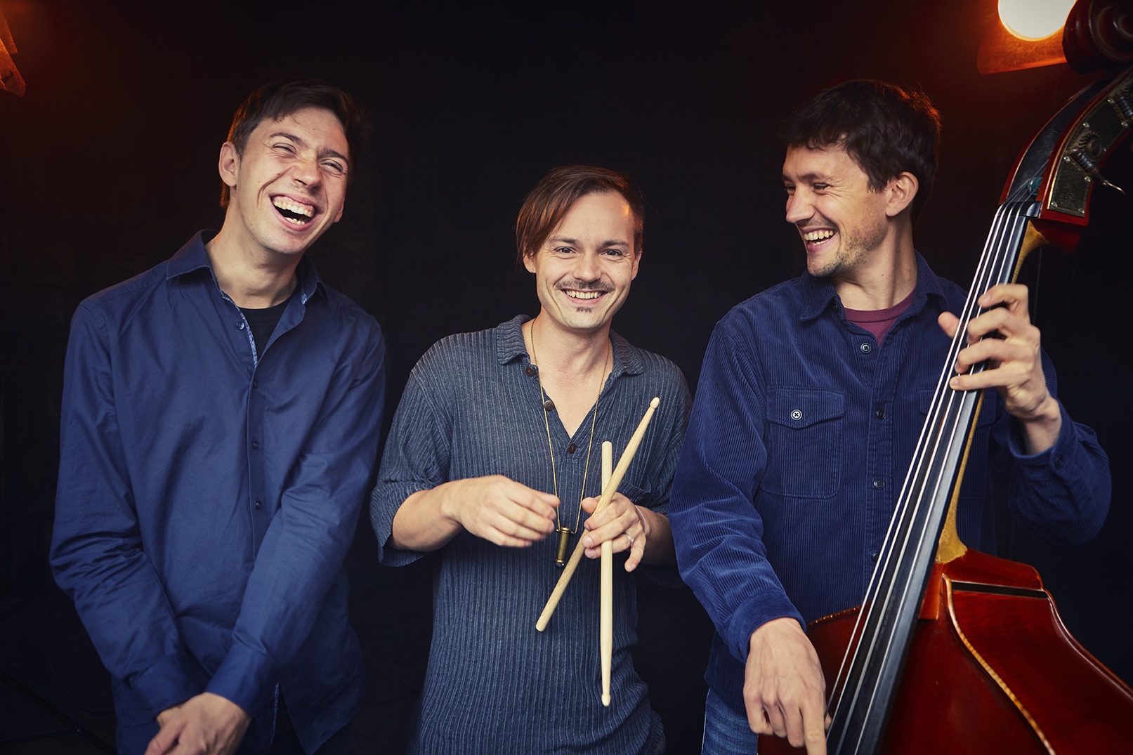Foto von drei Personen die lachen, eine hält einen Bass, eine andere Schlagzeugstöcke.
