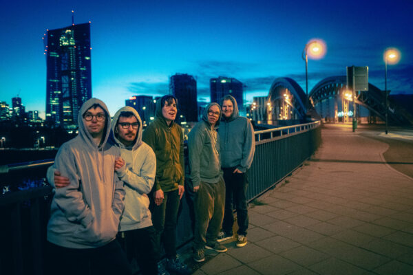 Buntfoto der fünf Mitglieder der Band auf einer Brücke, alle haben Hoodies an, im Hintergrund die Silhouette einer Stadt.