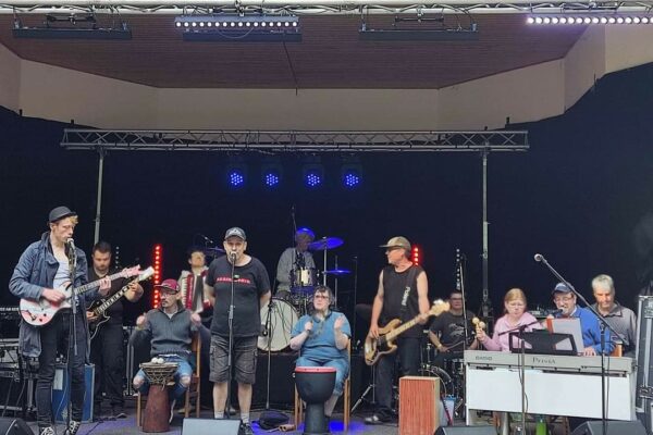Ein Foto von 12 Mitgliedern der Band bei einem Live-Auftritt auf einer großen Außenbühne.