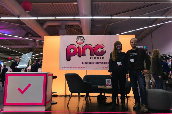 Katharina und Thorsten posieren am Pinc Music-Stand vor der Kamera. Am linken Bildrand steht ein pink-umrandeter Tresen mit einem Monitor. Im Hintergrund steht eine Couch, ein Sessel und ein Tisch. Dahinter hängt das Banner über einer orange-beleuchteten Wand.