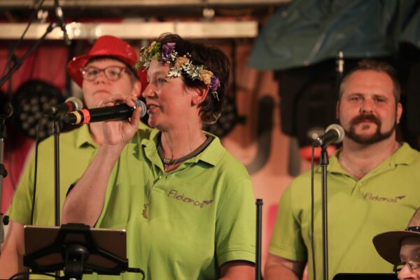 drei Mitglieder des Chors live auf der Bühne mit Mikrofonen. Alle tragen das gleiche Polo-Shirt.