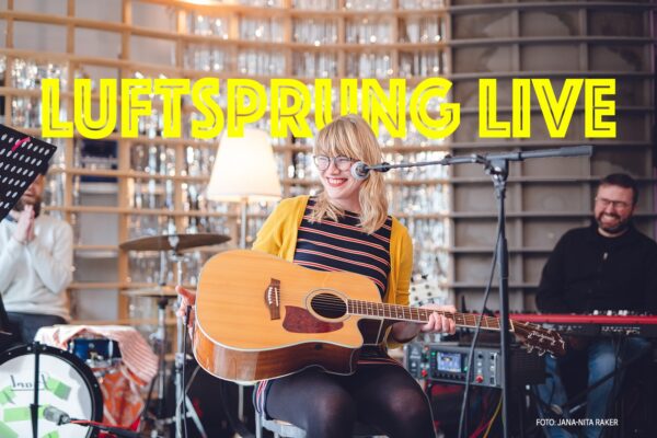 eine Frau mit Gitarre sitzt lachend mit Hocker vor Mikro auf der Bühne, im Hintergrund ein Musiker am Keyboard und einer am Schlagzeug, bei lachen