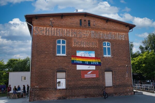 Buntfoto von einem Backsteingebäude mit zwei Stockwerken, daran eine Regenbogenfahne mit Schrift "Love is love" und ein Schild "Seebrücke Uckermark"