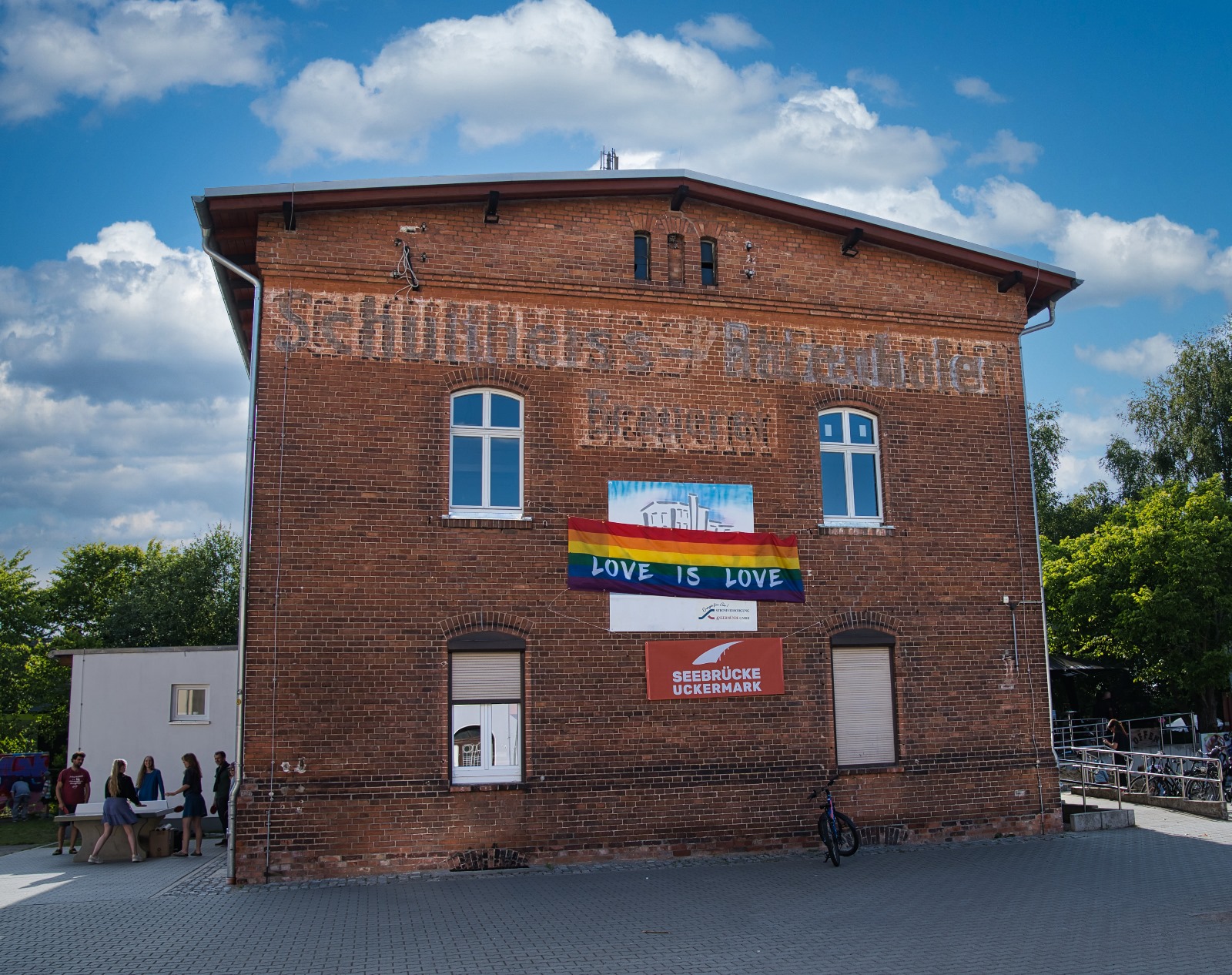 Buntfoto von einem Backsteingebäude mit zwei Stockwerken, daran eine Regenbogenfahne mit Schrift "Love is love" und ein Schild "Seebrücke Uckermark"