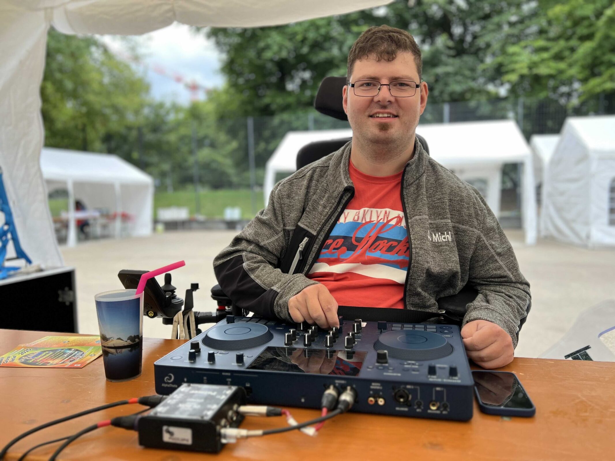 Ein Mann sitzt in einem Festivalzelt, vor ihm ein Tisch mit DJ Equipment , hinter ihm andere Felte und Bäume. Er sitzt am Mischpult in einem Rollstuhl.