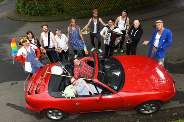 Foto von zwei Menschen in einem Cabriolet umringt von Menschen mit Staubwedeln und Musikinstrumenten