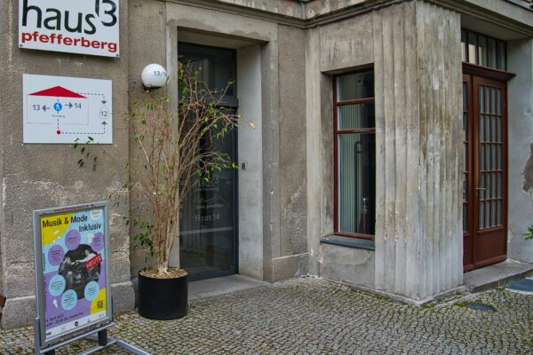 Buntfoto von einer Hausecke, mit einer großen Glastür, daneben ist ein Schild angebracht ,darauf das Logo des Pfefferberg haus13.