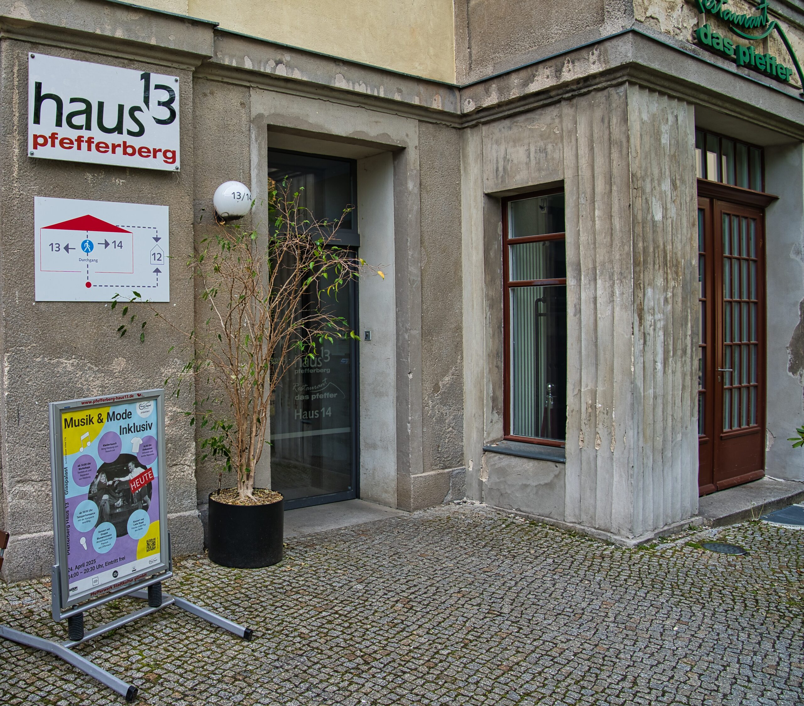 Buntfoto von einer Hausecke, mit einer großen Glastür, daneben ist ein Schild angebracht ,darauf das Logo des Pfefferberg haus13.