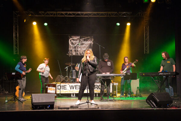 Buntfoto von einer Band mit sechs Mitglieder bei Auftritt, innen auf einer Bühne, mit Schriftzug DIE WEISSE ROSE