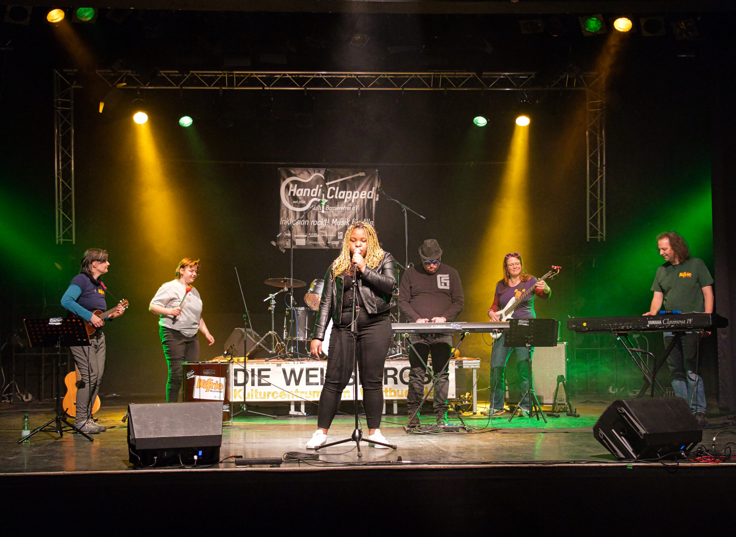 Buntfoto von einer Band mit sechs Mitglieder bei Auftritt, innen auf einer Bühne, mit Schriftzug DIE WEISSE ROSE
