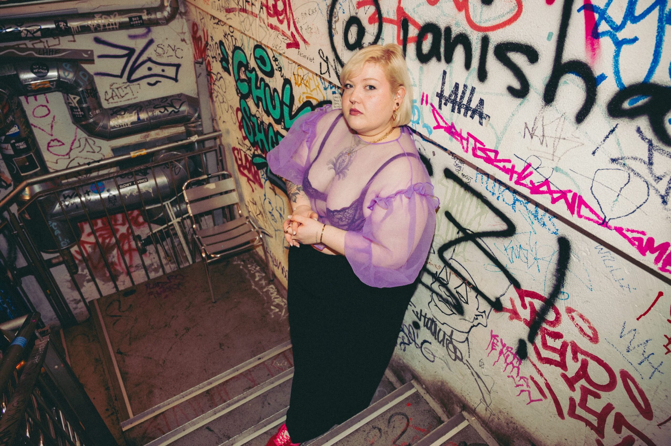 Bundfoto von einer Treppe, hinabblickend in ein Untergeschoß, an den Wänden vie Graffiti, in der Ecke ein Zwischenpodest auf dem ein einfacher Stuhl steht. An der Wand lehnt eine Frau. Sie trägt rote Turnschuhe, eine weite lange schwarze Hose, einen violetten BH und ein violett durchsichtiges Topp. Sie ist blond, trägt einen Nasenring und guckt hinauf in die Kamera.