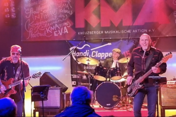 Bundfoto von drei Musikern live auf einer Bühne, innen. einer spielt Schlagzeug, einer E-Gitarre, einer E-Bass.