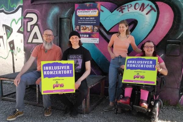 Vier Menschen alle sitzend, davon einer im Rollstuhl, sie halten zwei mit zwei großen "inklusiver Konzertort empfohlen von Pinc Music".