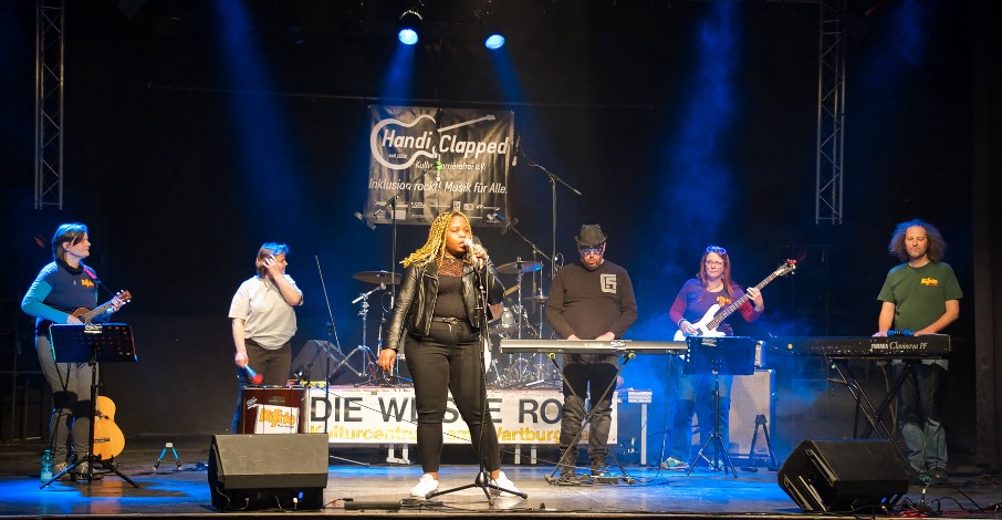 Foto von einer Band mit sechs Mitglieder bei Auftritt, innen auf einer Bühne, mit Schriftzug DIE WEISSE ROSE