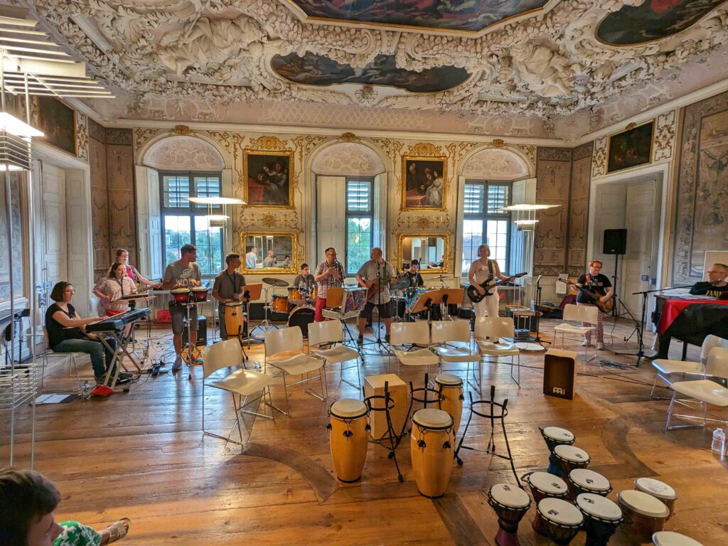 Buntfoto von einem tollen Saal in einem Schloss, mit prächtigem Stuck an der Decke und Gemälden an den Wänden. Darin stehen und sitzen 12 Musikerinnen und spielen Keyboards, Tongas, Schlagzeug, Gitarre, Bass und Klavier. Im Vordergrund stehen jede Menge Bongos mit denen Musiker:innen dazukommen könnten.