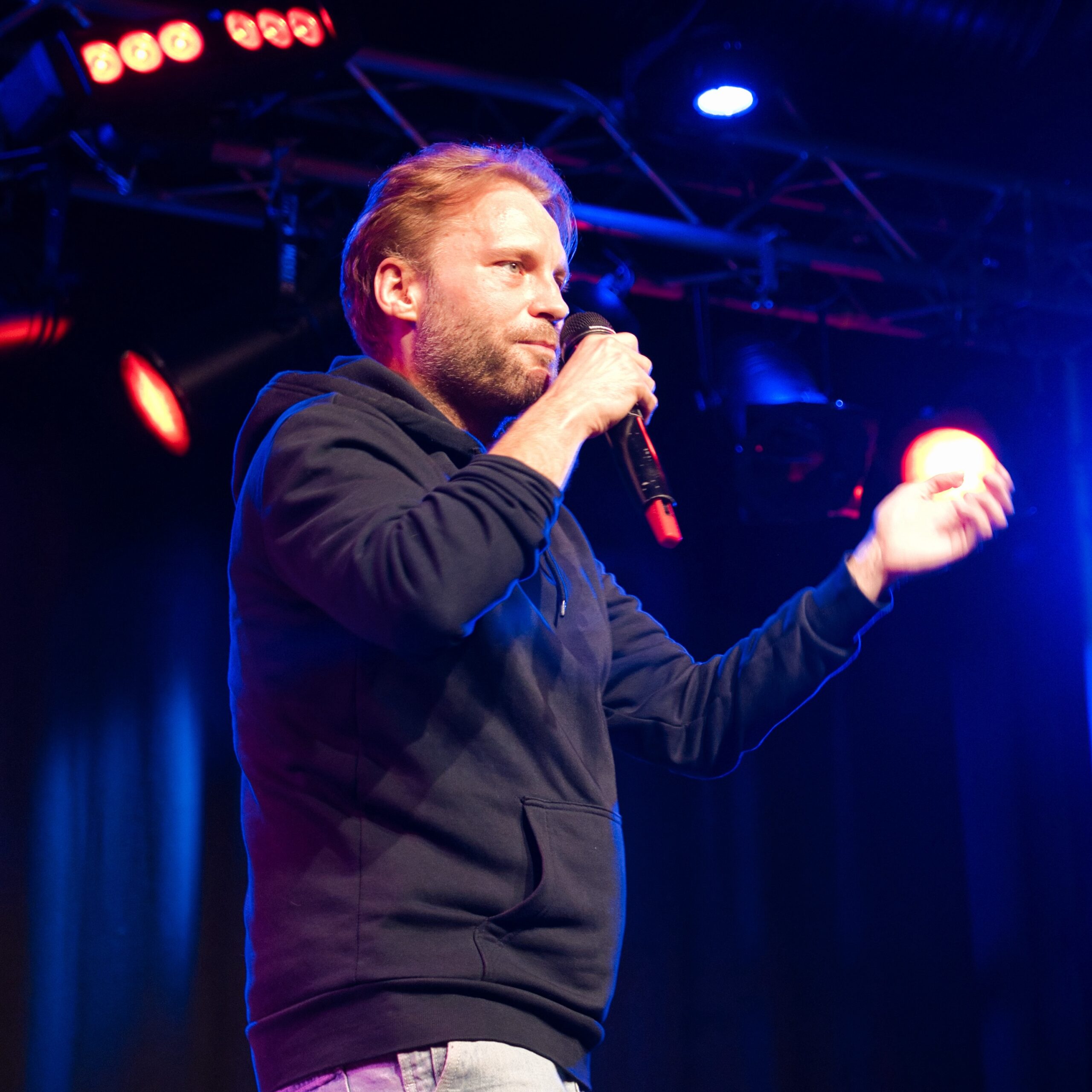 Ein junger Mann mit Pullover auf einer Bühne innen, der ins Mikrofon singt