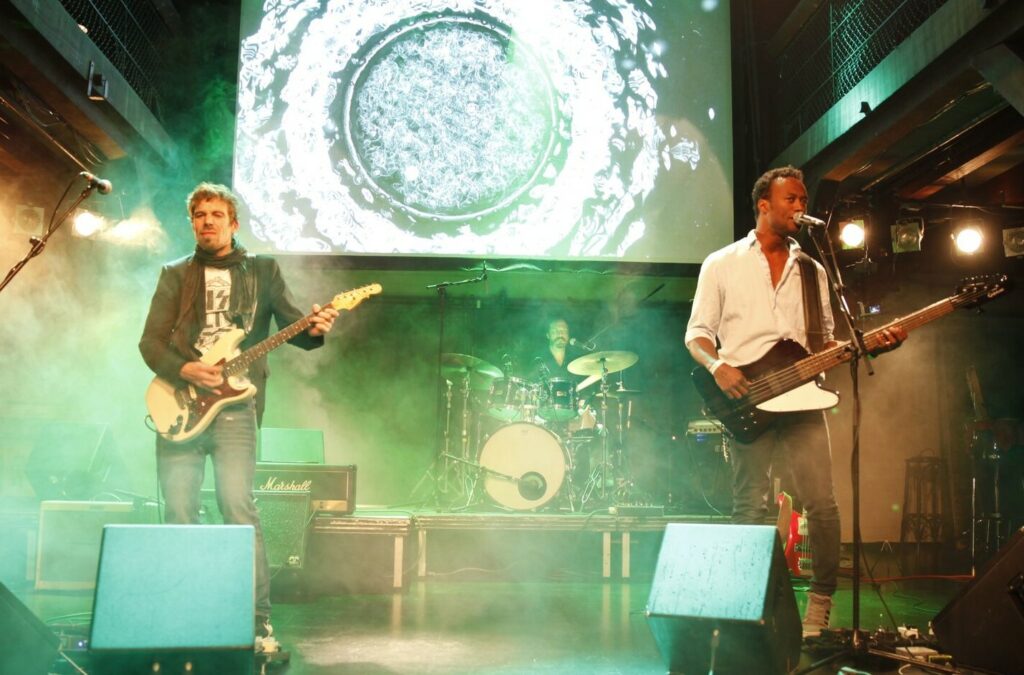 Drei Musiker mit Schlagzeug, E-Gitarre und Bass, im Hintergrund Videoleinwand und Bühnennebel.