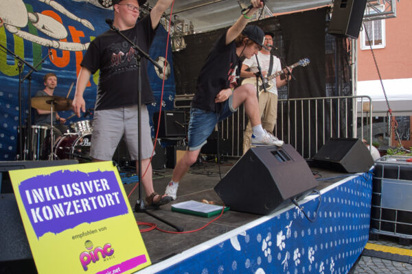 Buntfoto einer Band auf einer Außen-Bühne, im Hintergrund Rudolstadt Banner, im Vordergrund ein Schild "Inklusiver Konzertort" mit Pinc Msuzic Logo. Von der Band sind Schlagzeuger, Bassist und zwei Sänger zu sehen, letztere strecken die Mikros ins Publikum (dass sich außerhalb des Bildes befindet).