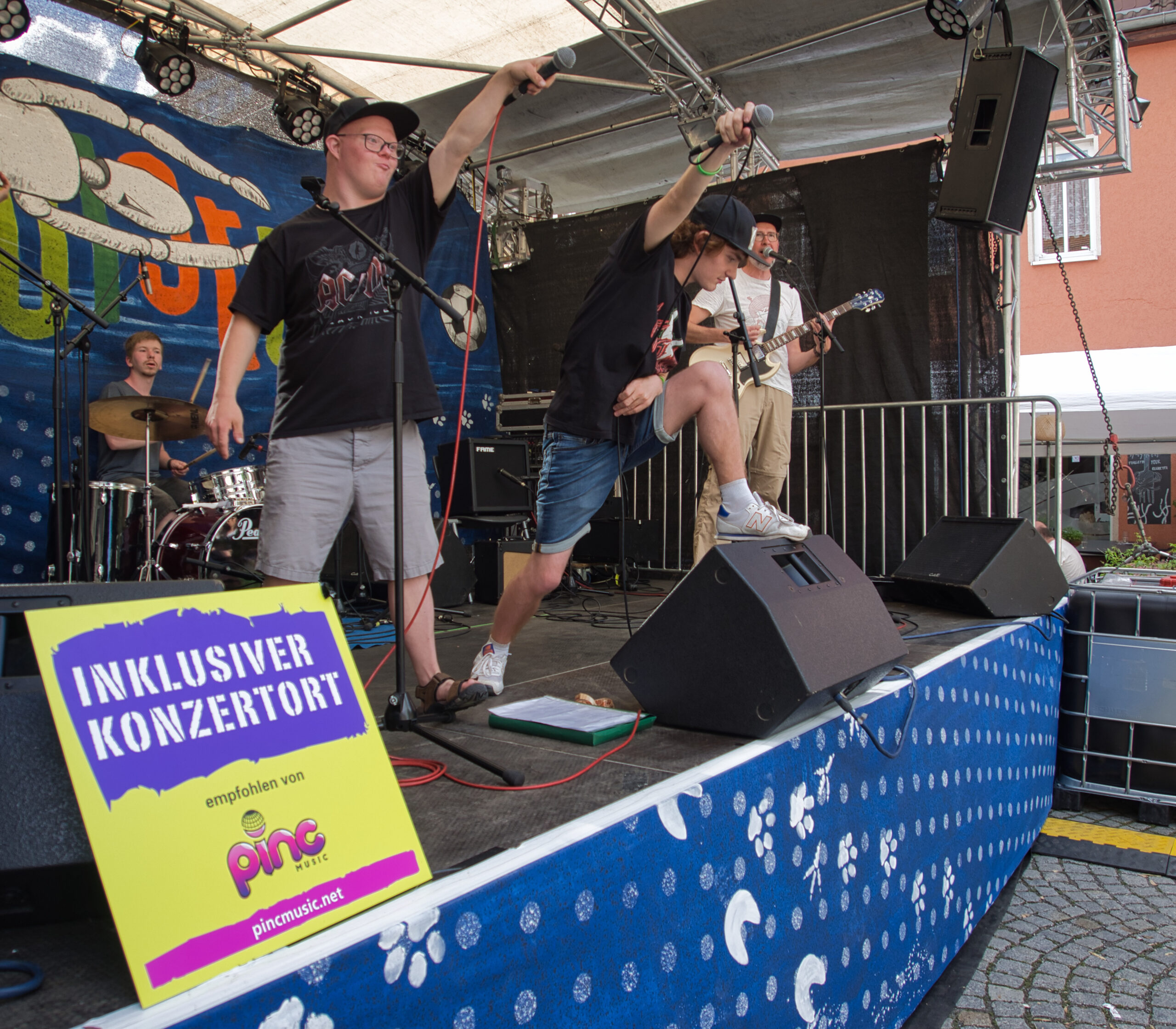 Buntfoto einer Band auf einer Außen-Bühne, im Hintergrund Rudolstadt Banner, im Vordergrund ein Schild "Inklusiver Konzertort" mit Pinc Msuzic Logo. Von der Band sind Schlagzeuger, Bassist und zwei Sänger zu sehen, letztere strecken die Mikros ins Publikum (dass sich außerhalb des Bildes befindet).