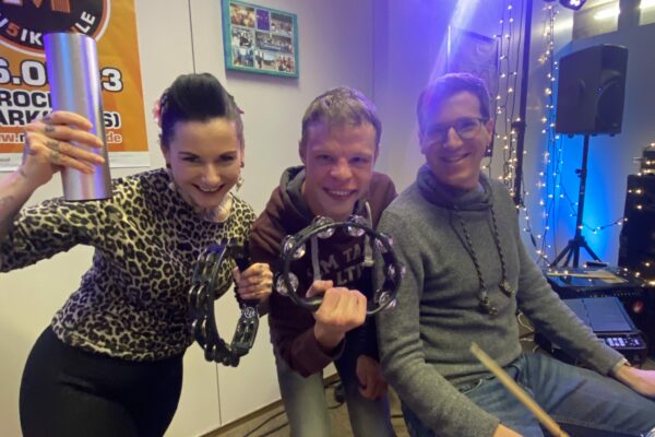 Buntes Foto von drei Menschen: zwei Mitgliedern der Band The Cool Chickpeas zusammenClaudia Lippmann vom Rundfunk-Tanzorchester Ehrenfeld - alle lächeln in die Kamera und haben Schlaginstrumente in der Hand.