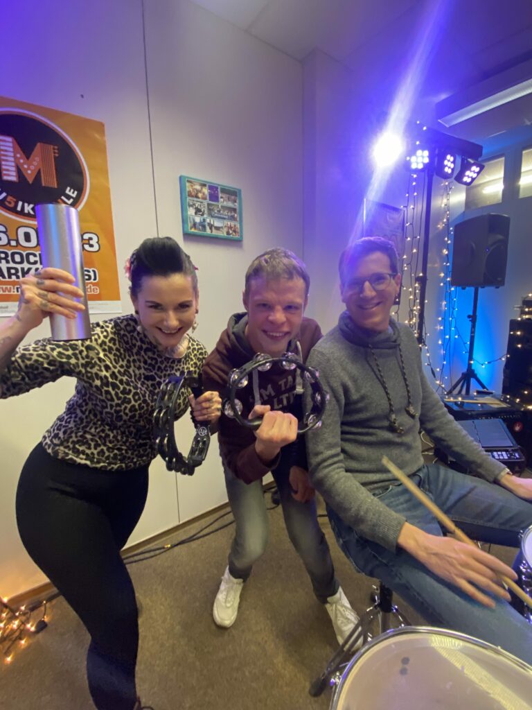 Buntes Foto von drei Menschen: zwei Mitgliedern der Band The Cool Chickpeas zusammenClaudia Lippmann vom Rundfunk-Tanzorchester Ehrenfeld - alle lächeln in die Kamera und haben Schlaginstrumente in der Hand.