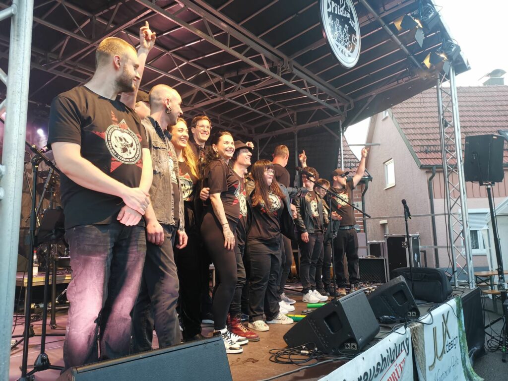 Buntes Foto von der Band The Cool Chickpeas zusammenClaudia Lippmann vom Rundfunk-Tanzorchester Ehrenfeld und Vom Ritchie von den Toten Hosen. Alle stehen zum Verbeugen gemeinsam auf einer Außenbühne. Von der Seite fotografiert.
