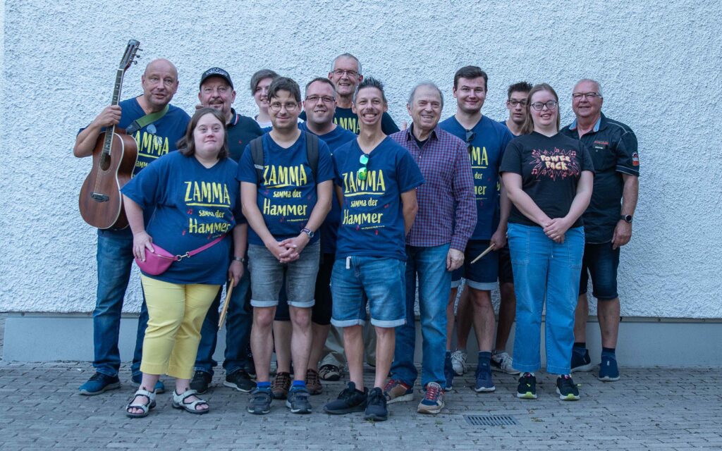 Buntes Gruppenfoto mit elf Mitgliedern der Band PowerPack und Günter Sigle und Barney von der Spider Murphy Gang. Alle stehen gemeinsam vor einer weißen Wand und lächeln in die Kamera.