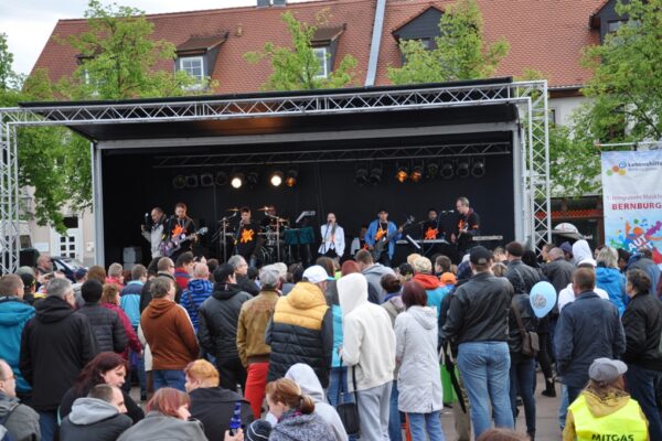 9 Mitglieder einer Band beim Live Auftritt auf einer Außenbühne mit viel Publikum dafür (über 100 Menschen). Sie speilen verschiedene Instrumente, u.a. E-Bass, E-Gitarre und Schlagzeug.