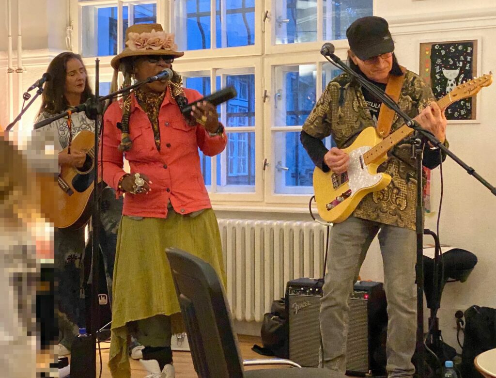 Bundfoto von zwei Frauen und einem Mann live beim Auftritt in einem Raum. Eine Frau spiel Akustikgitarre, eine Frau singe, der Mann spiel E-Gitarre.