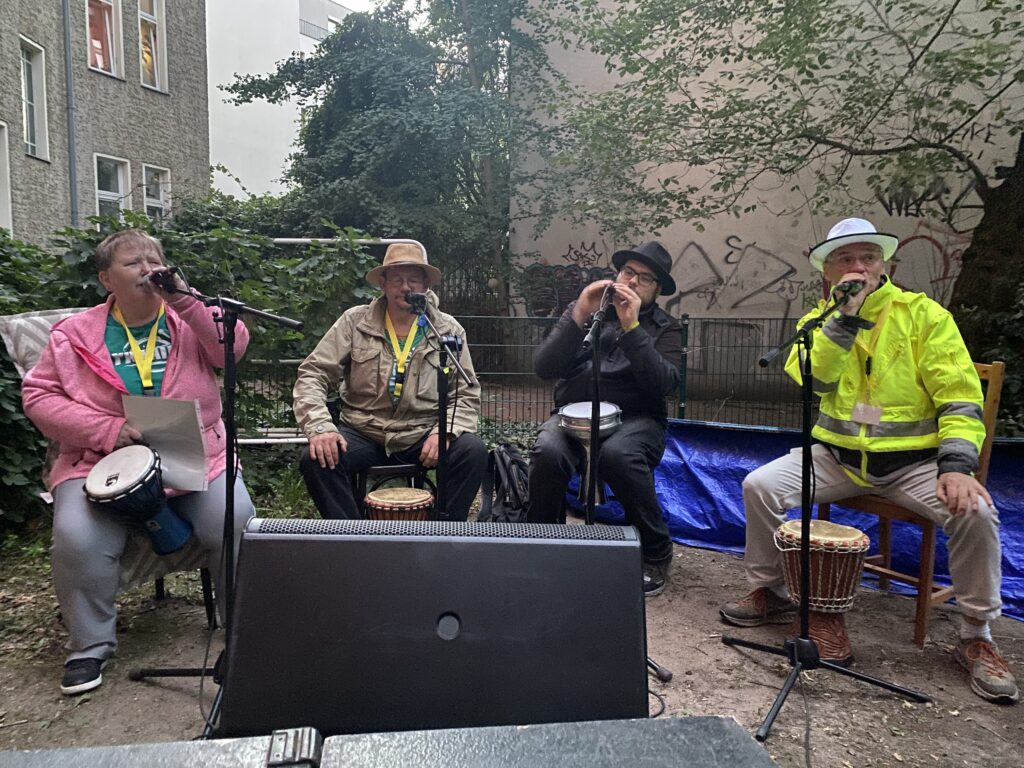 Buntfoto von vier Mensen, die mit Bongos auf Stühlen sitzen und in Mikrofone singen. Drei tragen Hüte. Sie sitzen in einem Innenhof in der Stadt mit zwei Bäumen im Hintergrund.