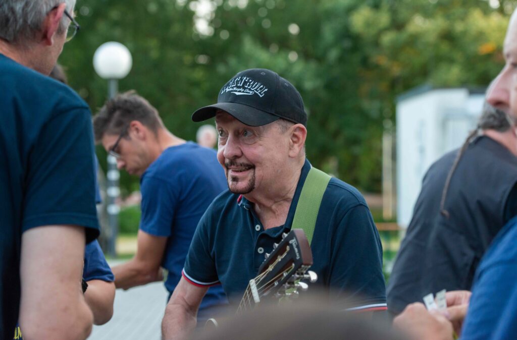 Buntfoto: Barny von der Spider Murphy Gang spielt Gitarre, um ihn rum unscharf im Anschnitt Mitglieder der Band PowerPack