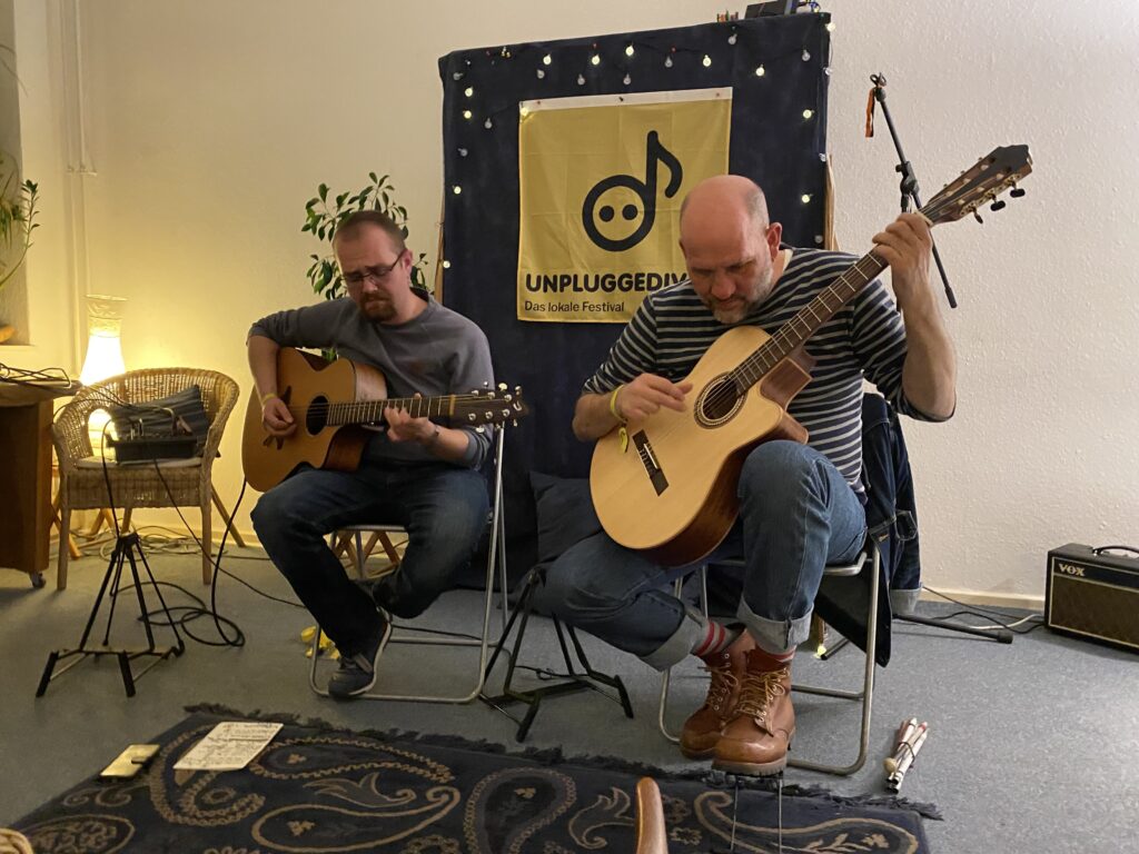 Buntfoto von zwei Musikern mit Gitarren, die nebeneinander sitzend ein Konzert geben, innen. Im Hintergrund ein Plakat mit dem Unpluggedival Logo und Schriftzug.